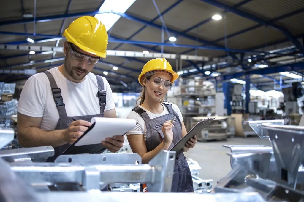 Due tecnici con casco giallo e occhiali di sicurezza controllano appunti in officina.